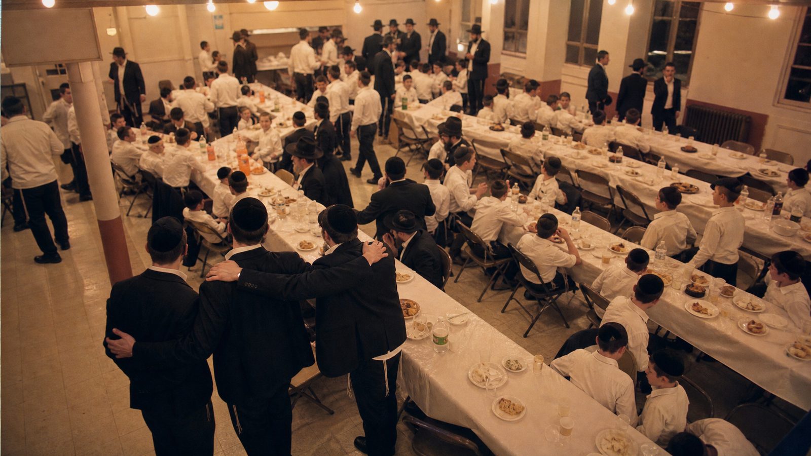 A siyum in a school social hall, long tables and crowded benches